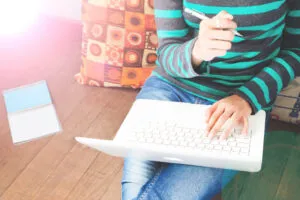 A person in a striped shirt and jeans sits on the floor with a laptop, looking ready to write for humans' search intent, holding a pen with a notebook nearby and colorful cushions behind them.